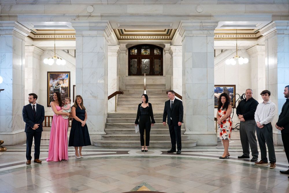 Wedding party and guests stand in semicircle before grand marble staircase in ornate courthouse rotunda for ceremony.