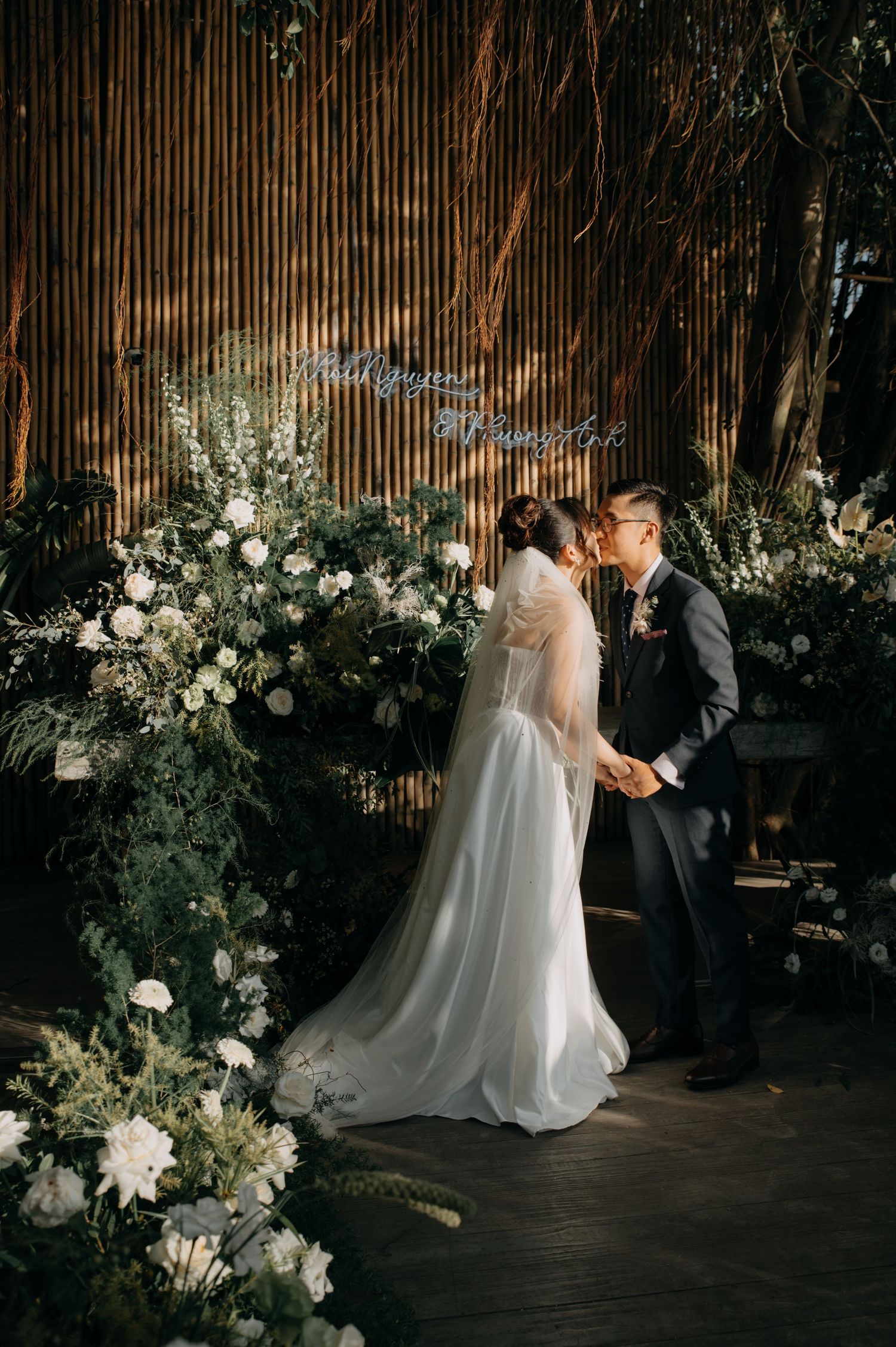 Romantic wedding kiss framed by lush white florals against a rustic bamboo backdrop.