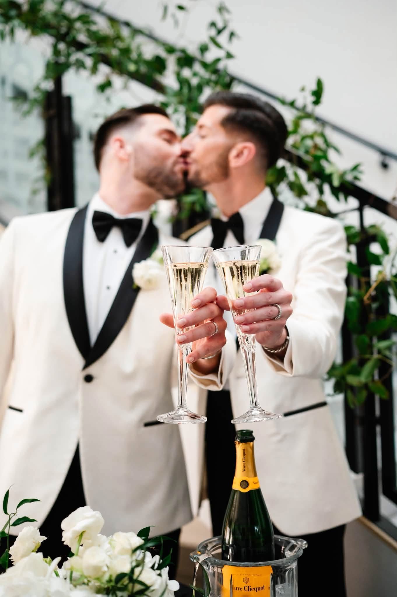 Two grooms in white tuxedos share a kiss while toasting with champagne flutes at their wedding celebration.