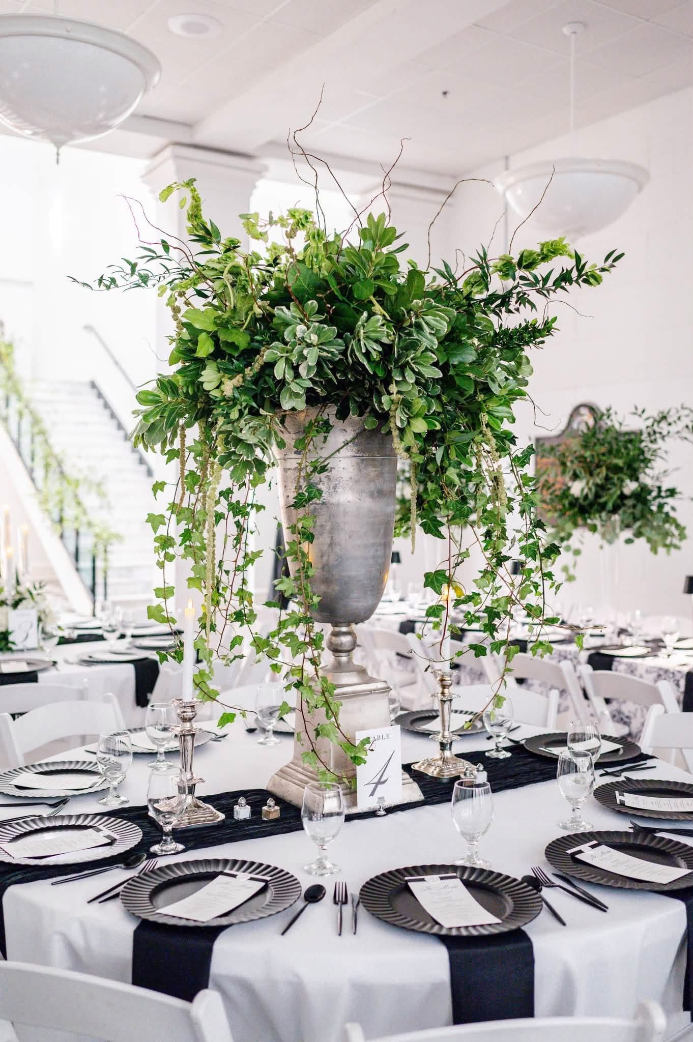 Elegant wedding reception table with tall silver urn centerpiece overflowing with lush greenery, black napkins, and silver charger plates.