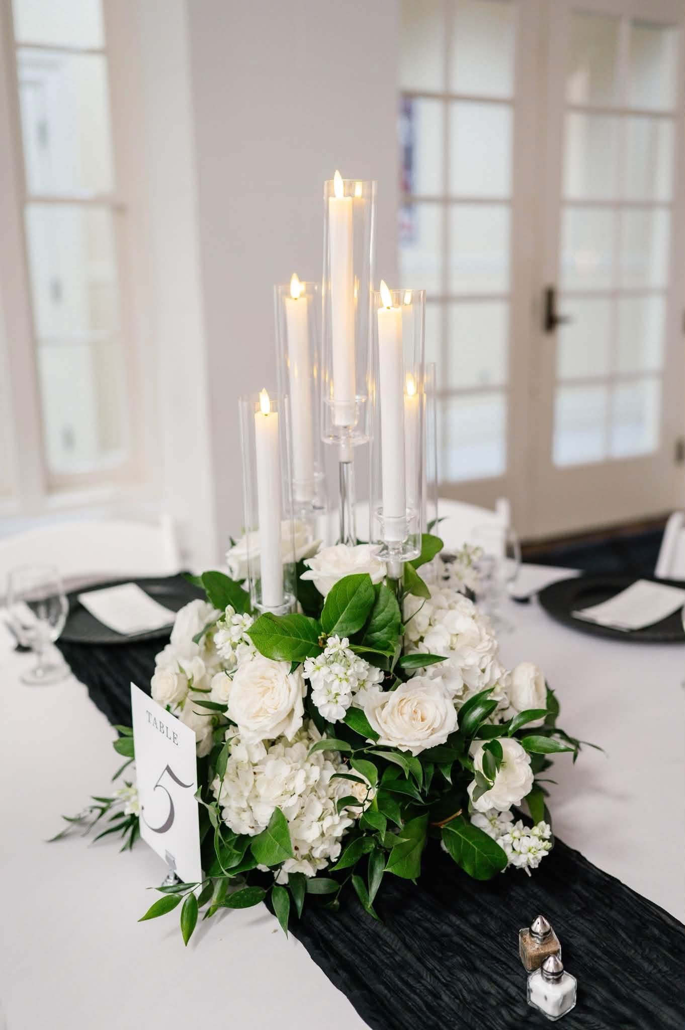 Elegant wedding centerpiece with white candelabra, white roses, hydrangeas, and greenery on a black table runner.