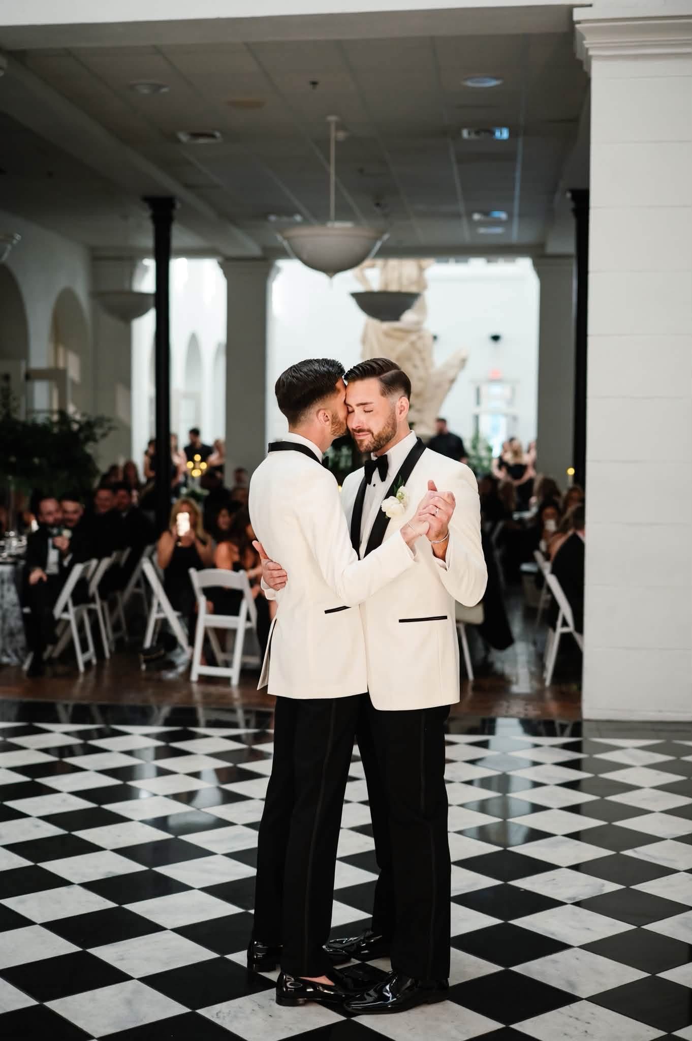 Two grooms share their first dance in matching white tuxedo jackets on a black and white checkered dance floor.