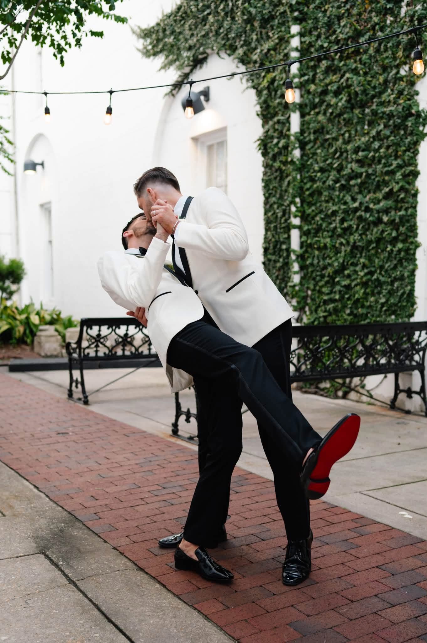 Two grooms in white tuxedo jackets share a romantic dip pose on a brick courtyard surrounded by ivy walls.