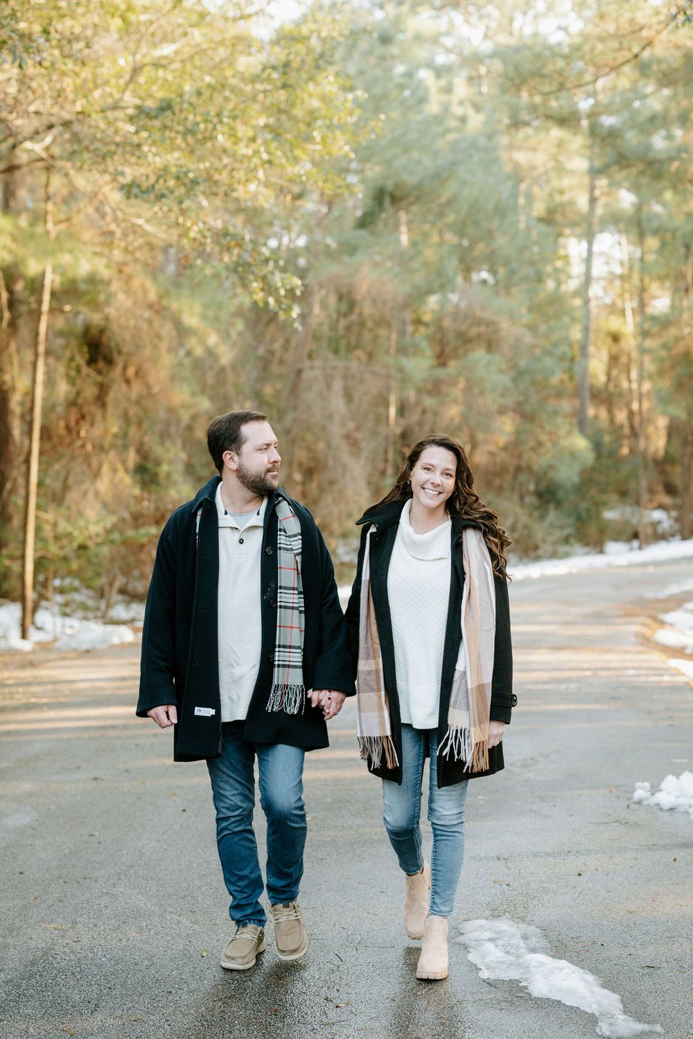 A couple walks together on a snowy path through pine trees while wearing winter coats and scarves.