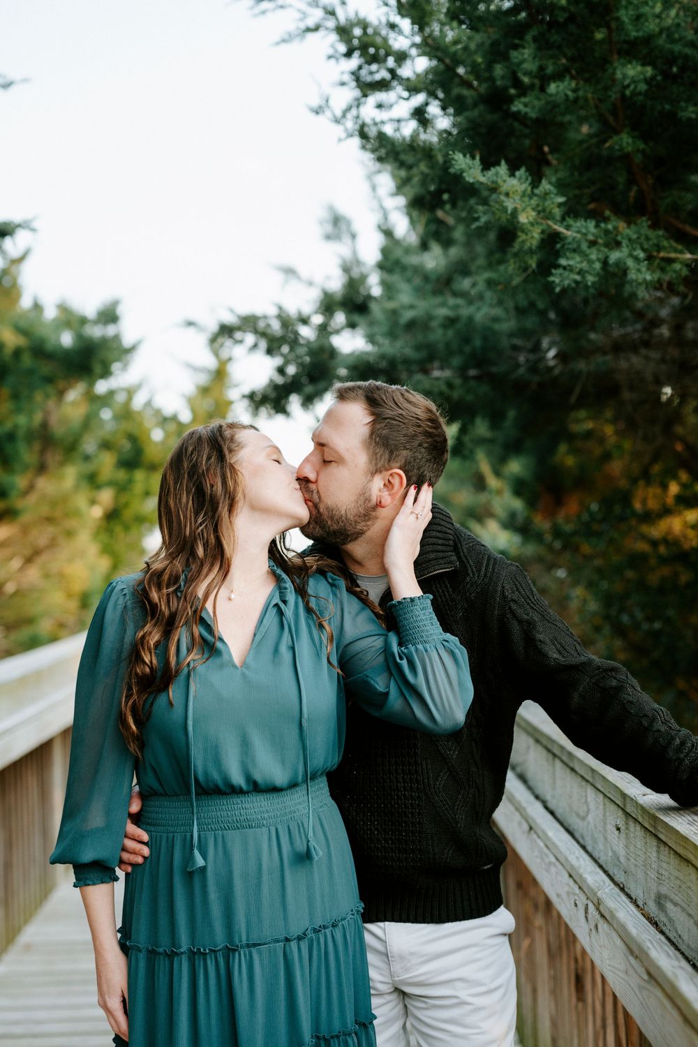 A couple shares an intimate kiss while standing on a wooden deck surrounded by evergreen trees.