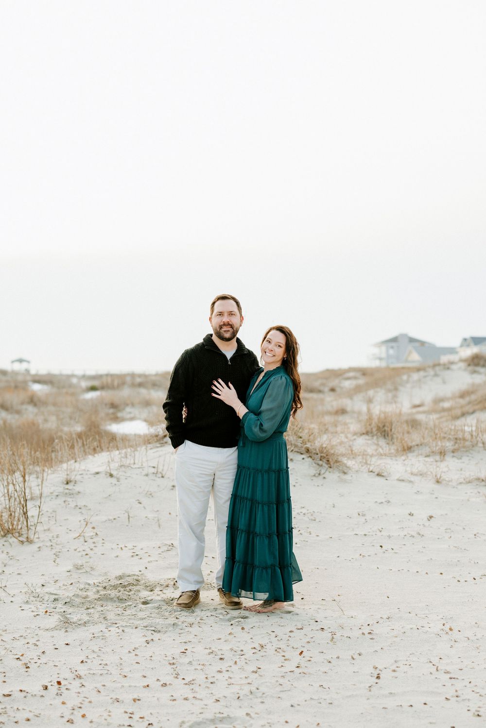 A couple poses together on sandy dunes wearing coordinated outfits of black and teal during a beach photoshoot.