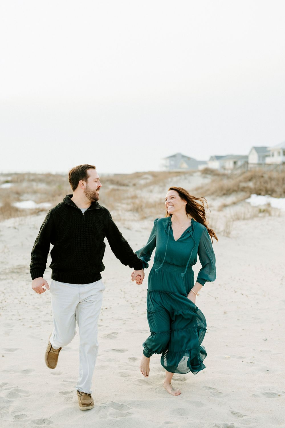 A couple runs playfully along the beach sand while wearing casual outfits in black and teal colors.