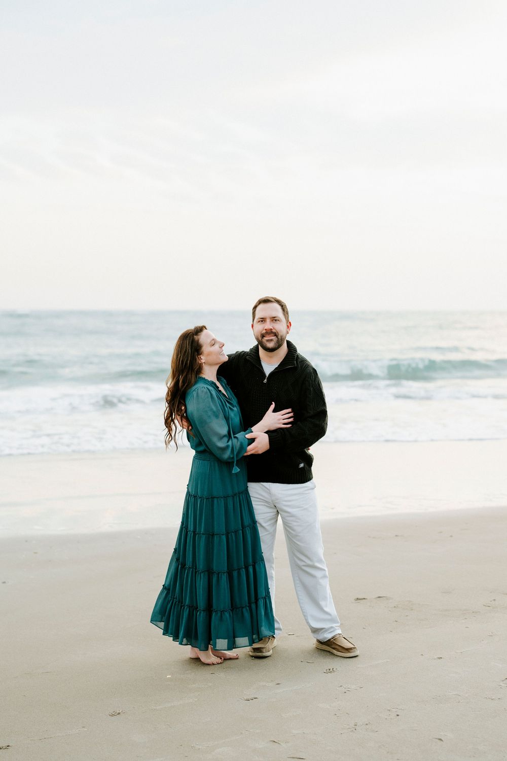 A couple shares joyful moments together during a beach photoshoot in teal dress and casual white attire.
