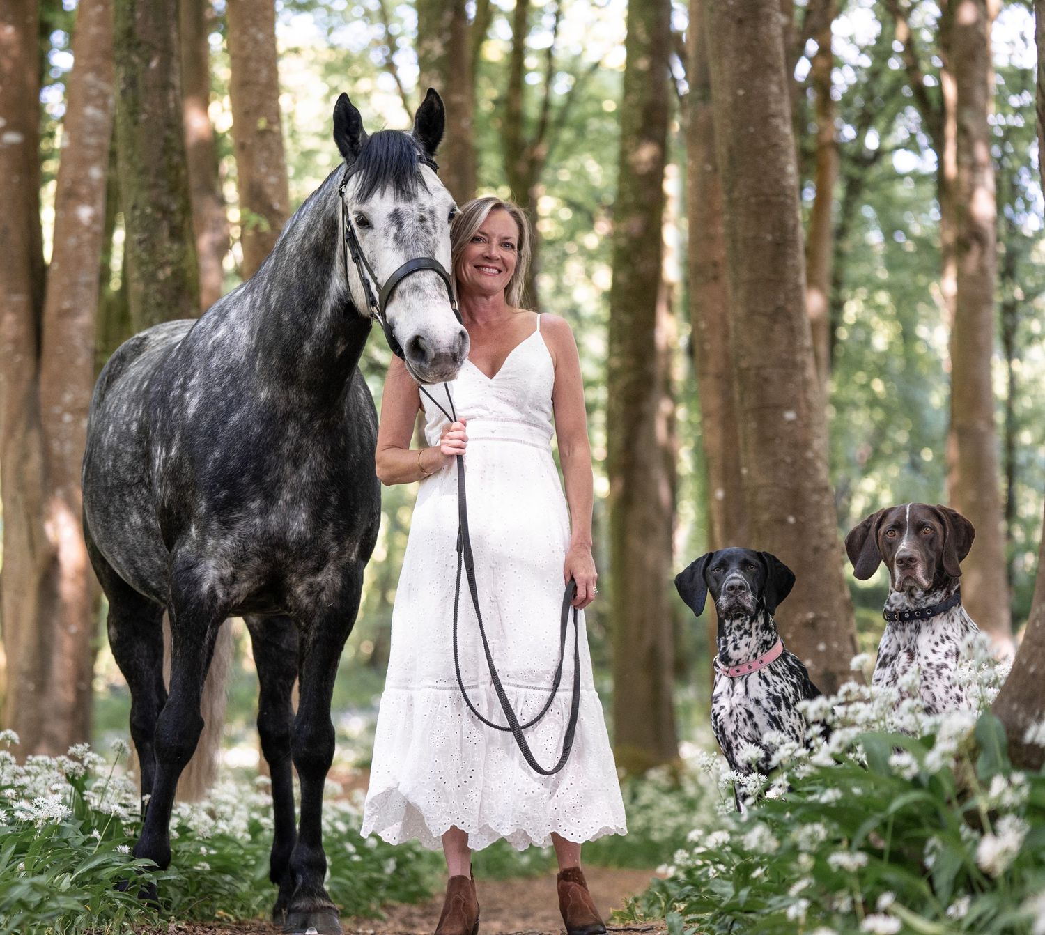 A dappled grey horse and two spotted dogs pose with a person in a white dress in a wooded forest setting.