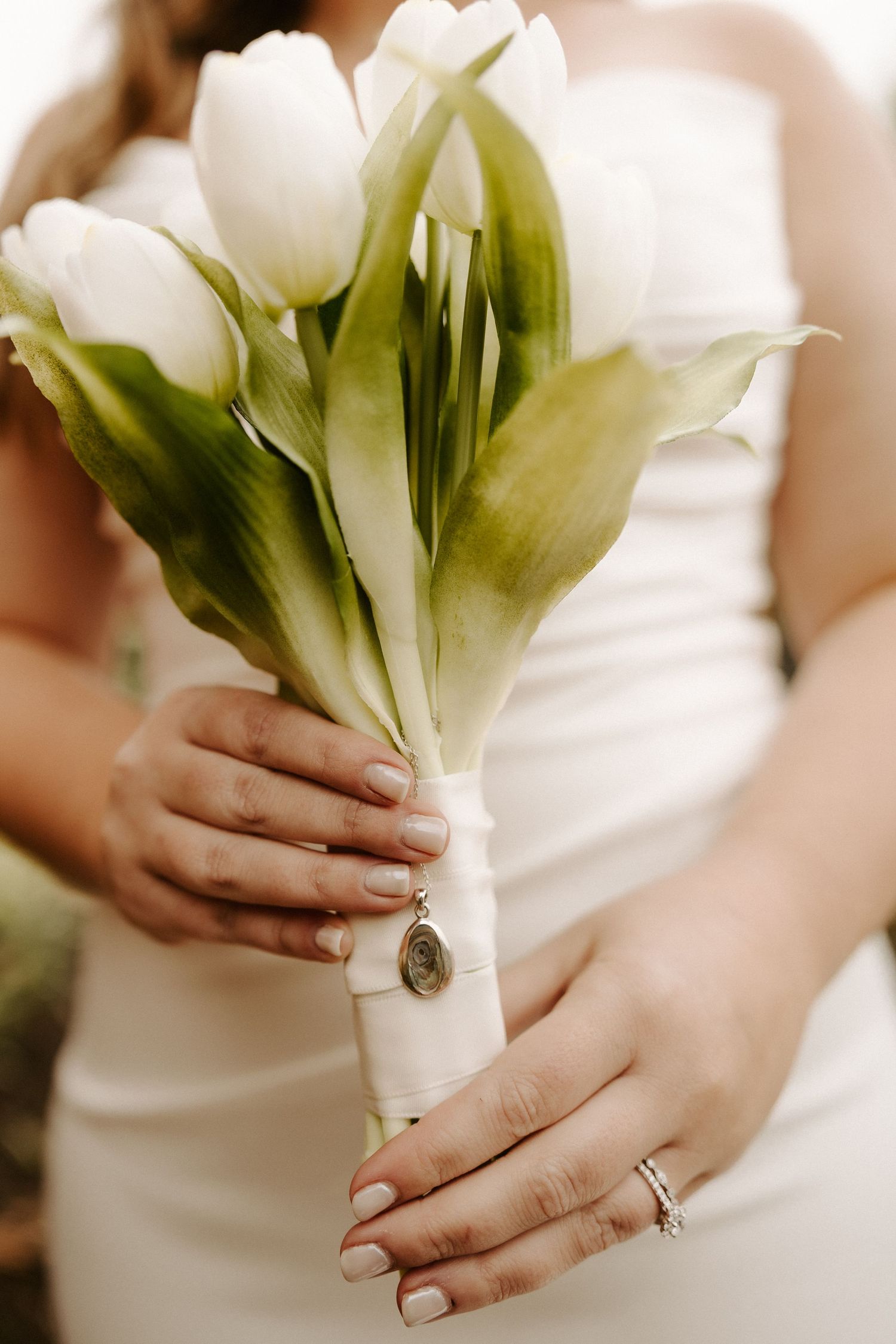 Bride holds a simple white tulip bouquet wrapped in white ribbon with a necklace attached.