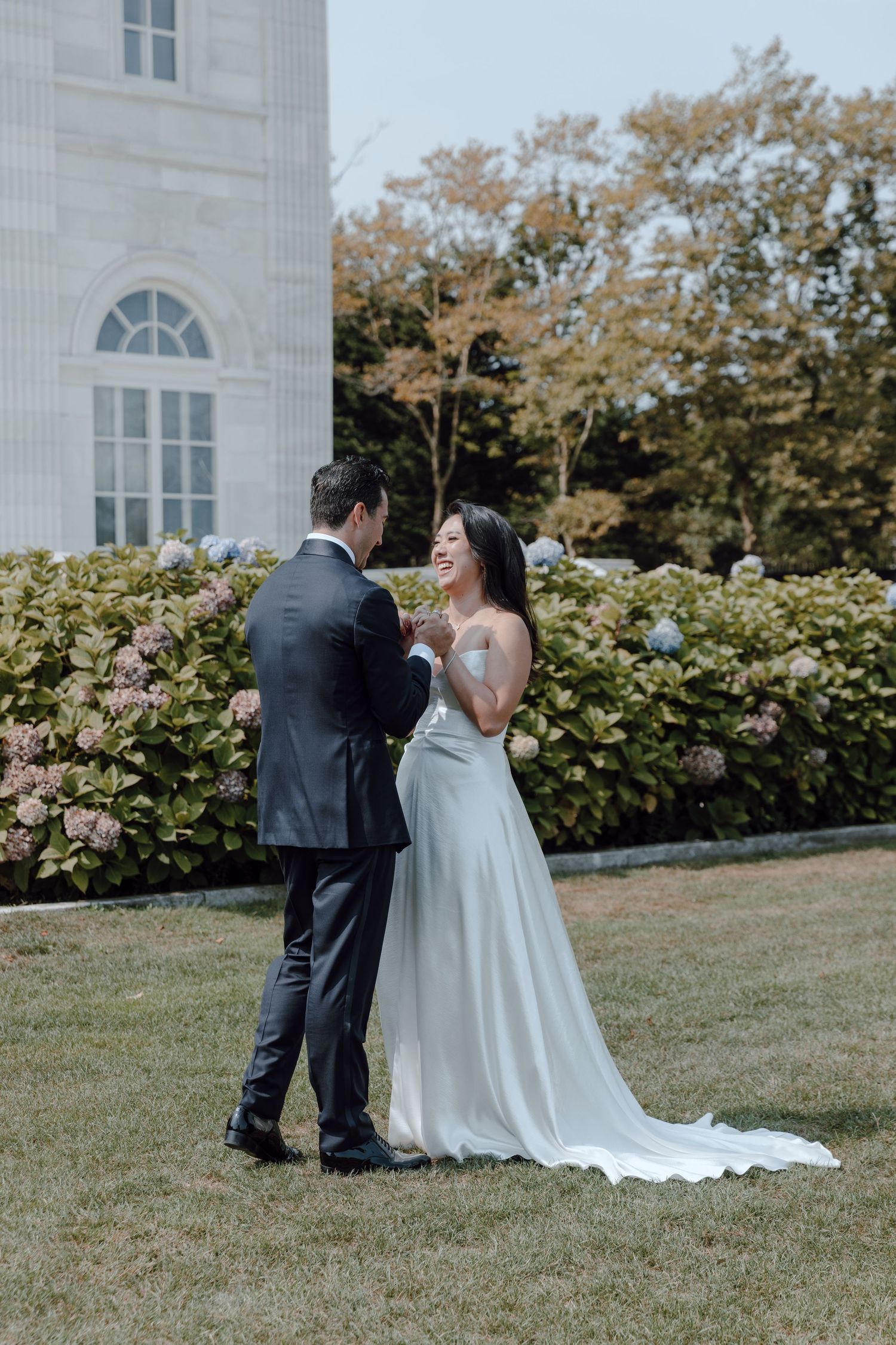 A couple shares a romantic moment on their wedding day on a manicured lawn beside a white building.