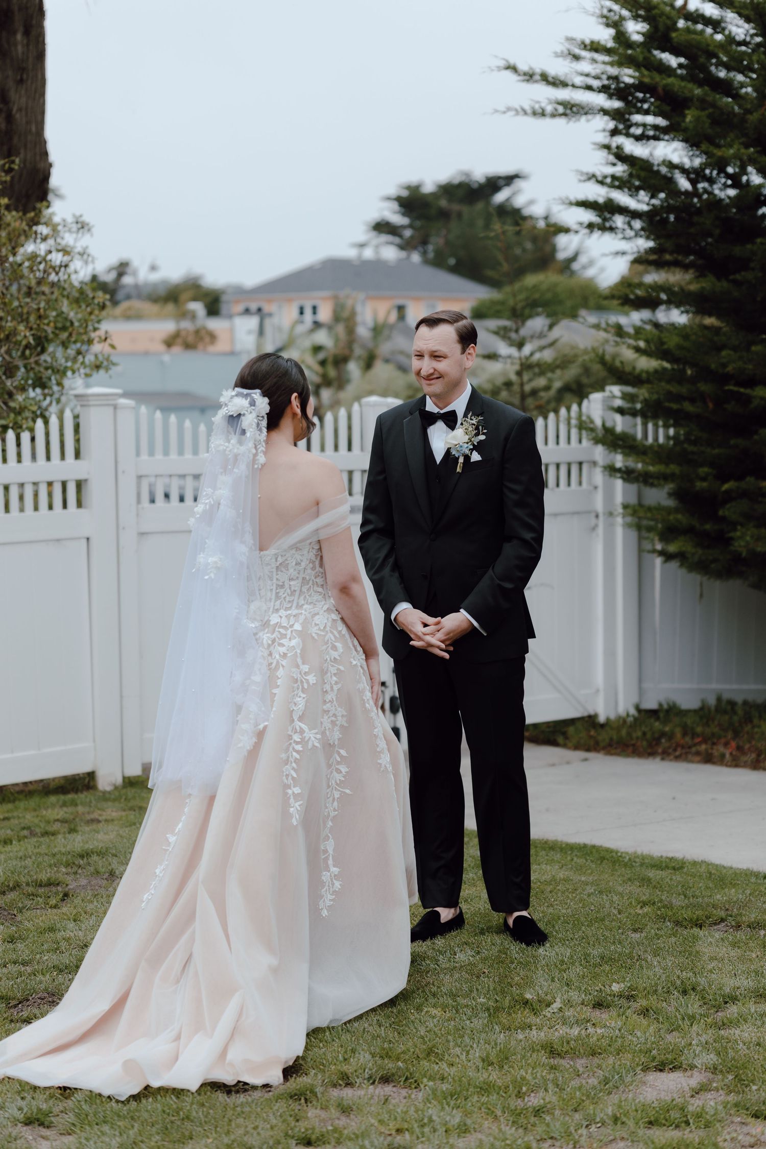 A bride and groom share a first look moment outdoors by a white picket fence on their wedding day.