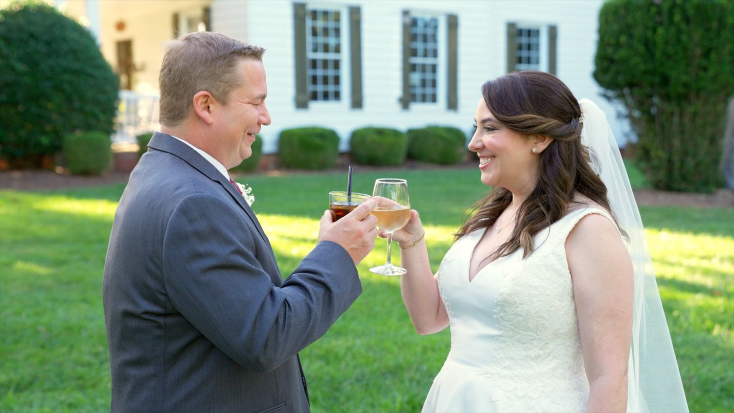 A couple in wedding attire shares a celebratory toast outdoors on a sunny day near a white building.