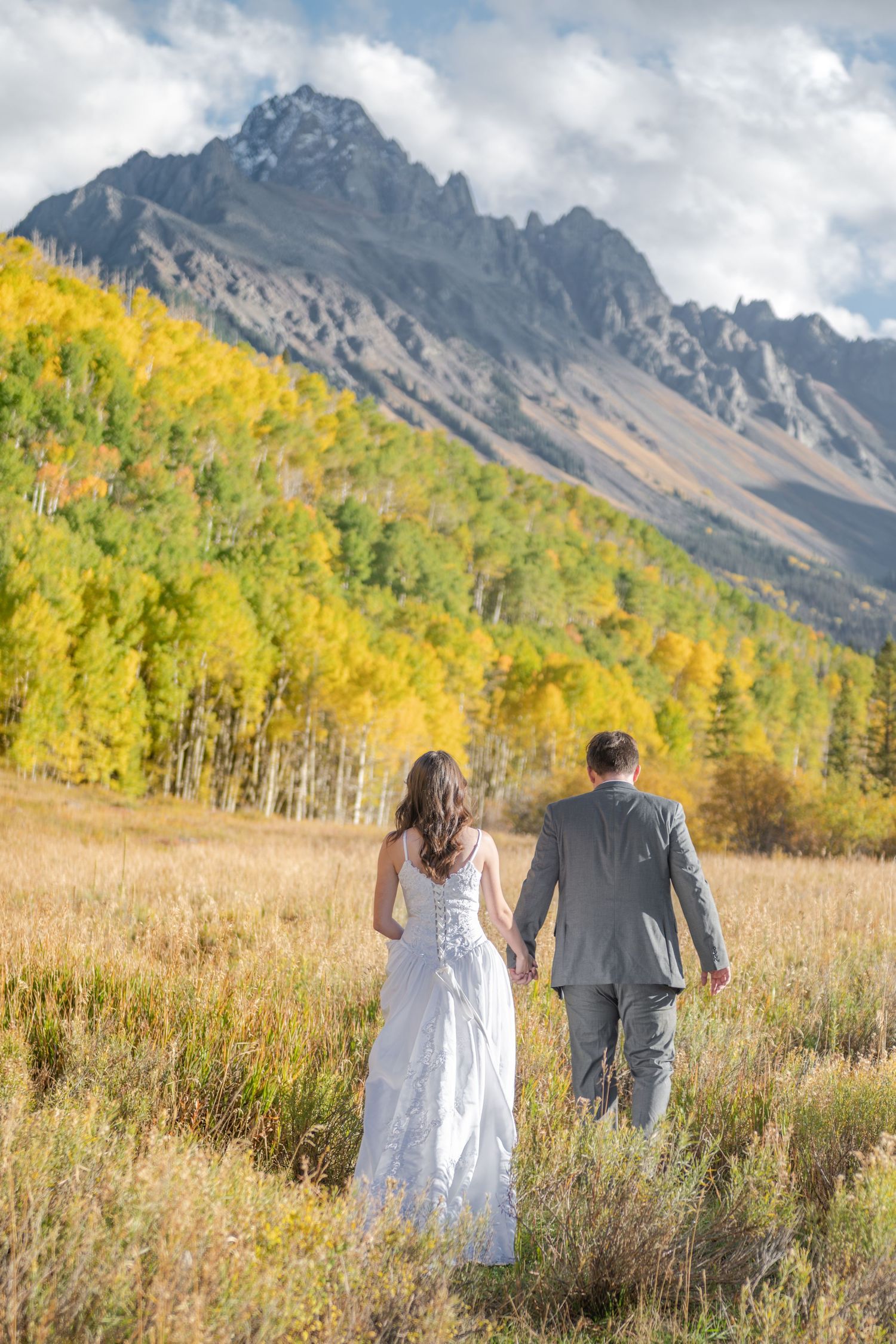An eloping couple walks hand in hand through a golden autumn meadow with majestic peaks and yellow aspen trees in the background.