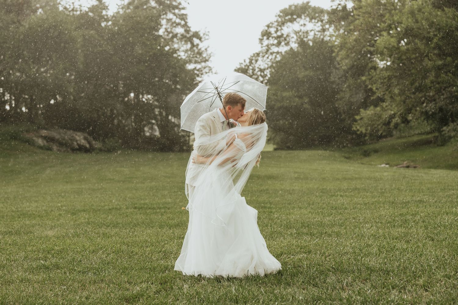 A romantic wedding couple embraces in a sunlit meadow while the bride's white dress flows in the breeze.