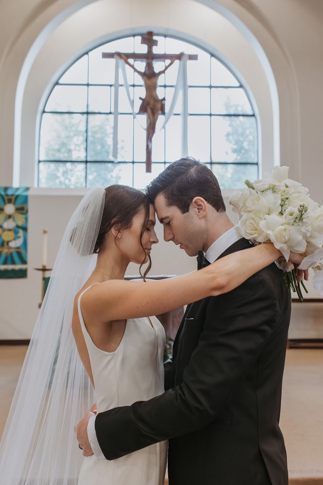 Romantic wedding couple shares intimate moment in front of arched church windows with natural light streaming in.
