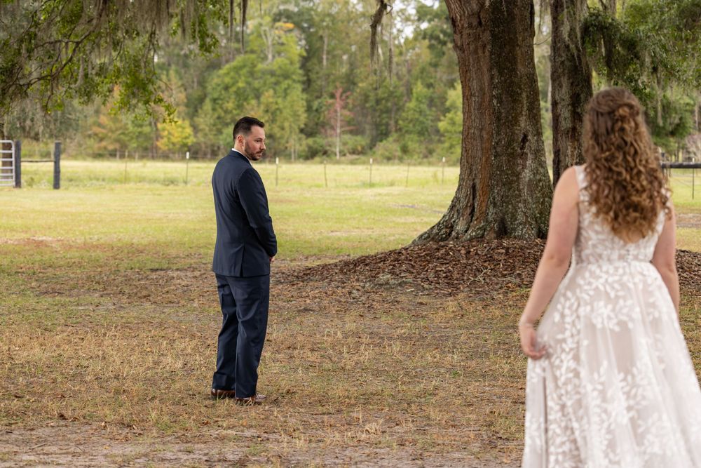 A couple stands apart during their first look moment under a large oak tree in a serene outdoor setting.