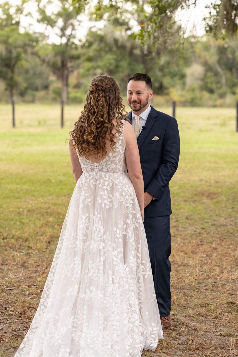 A couple shares a romantic first look moment in an outdoor setting wearing a white lace gown and navy suit.