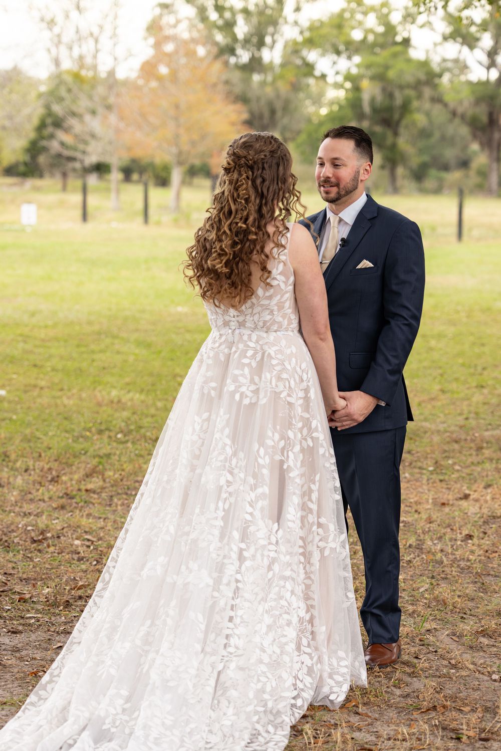 A couple in wedding attire stands together in a grassy field during autumn, with the bride wearing a flowing lace dress.