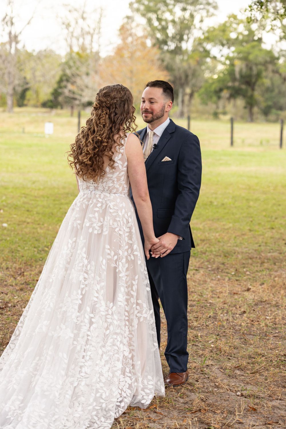 A couple in wedding attire stands together in a grassy field during autumn.
