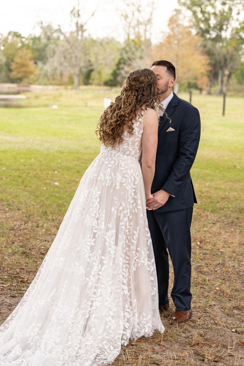 A romantic wedding moment captured outdoors with a bride in a lace gown and groom in navy suit sharing an intimate embrace.