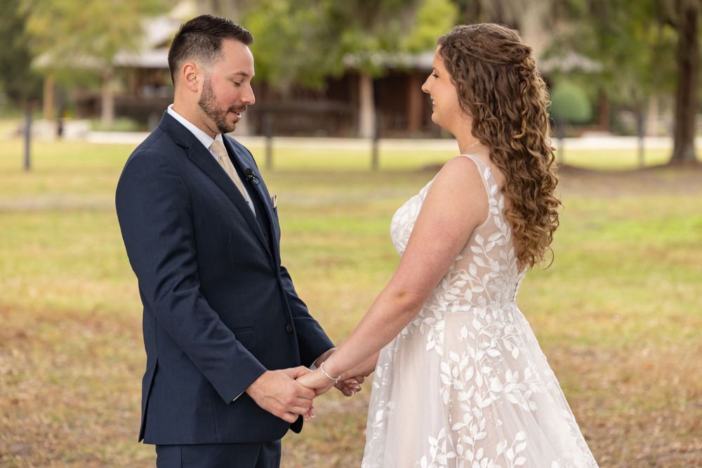 A couple shares a romantic moment outdoors during their wedding, wearing a navy suit and white lace gown.
