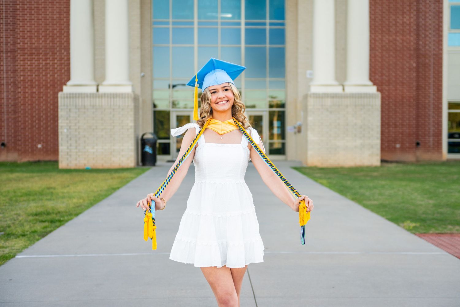 A person in a white dress twirls playfully on a concrete path between grassy areas and brick buildings.