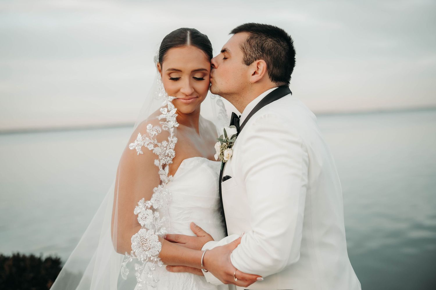 Groom kissing bride on cheek outdoors with ocean view, bride wearing lace veil and strapless gown.