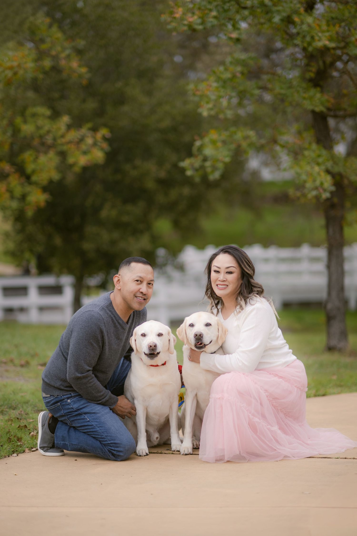 A couple sits with their two happy white dogs on a path surrounded by autumn trees.