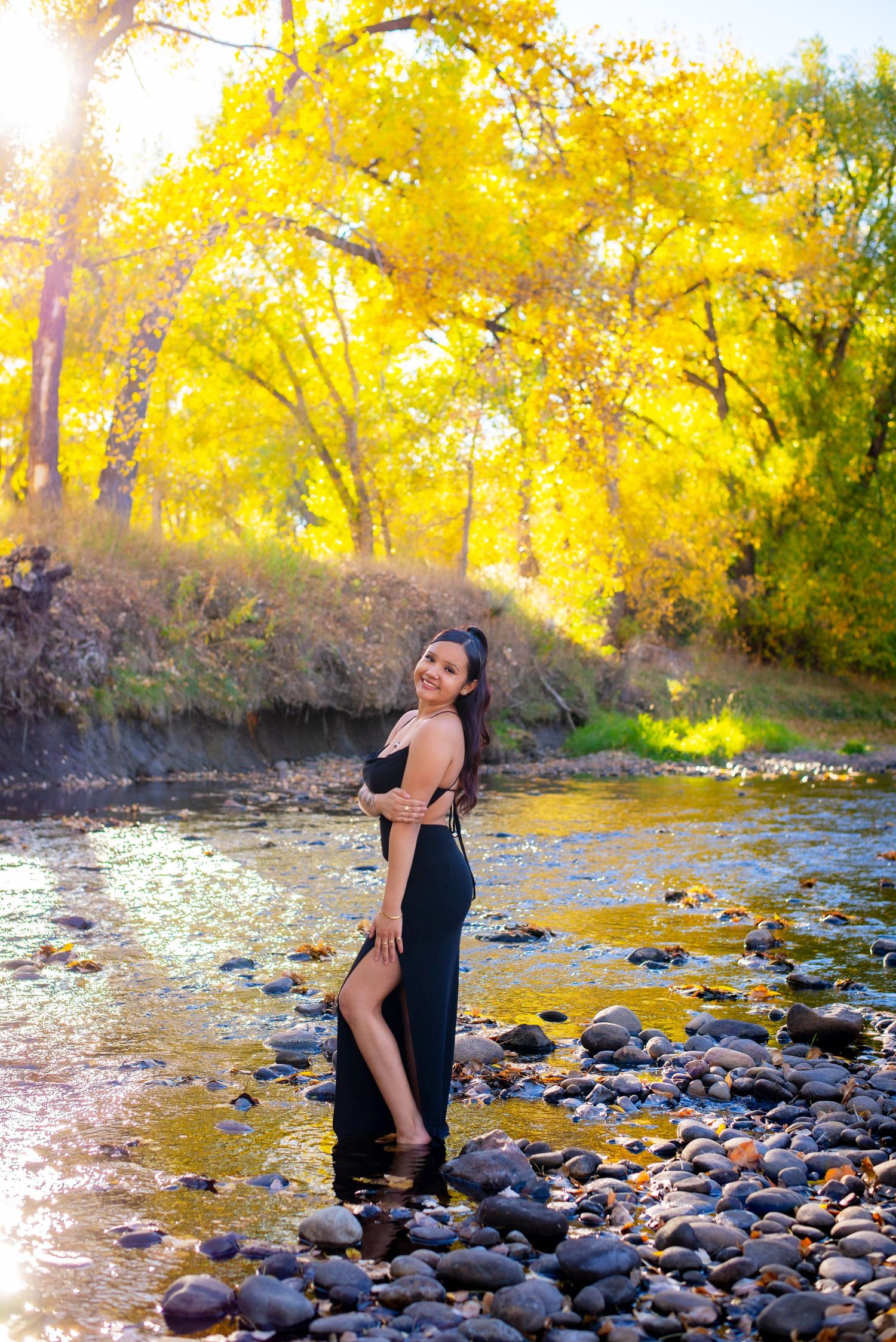 Woman in black dress poses by rocky stream with autumn foliage backdrop.