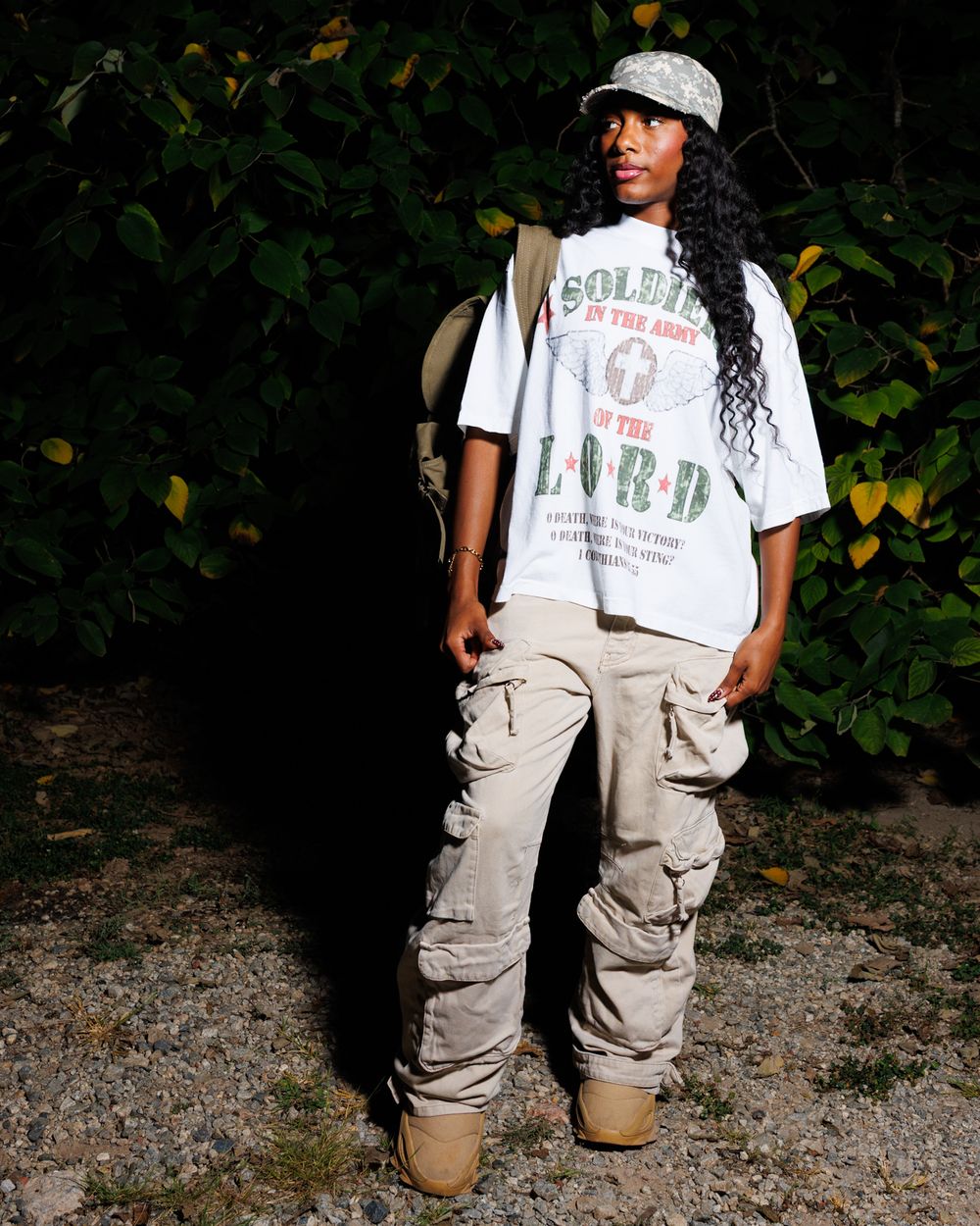 Person wearing a white graphic t-shirt and khaki cargo pants poses casually in an outdoor setting with foliage in background.