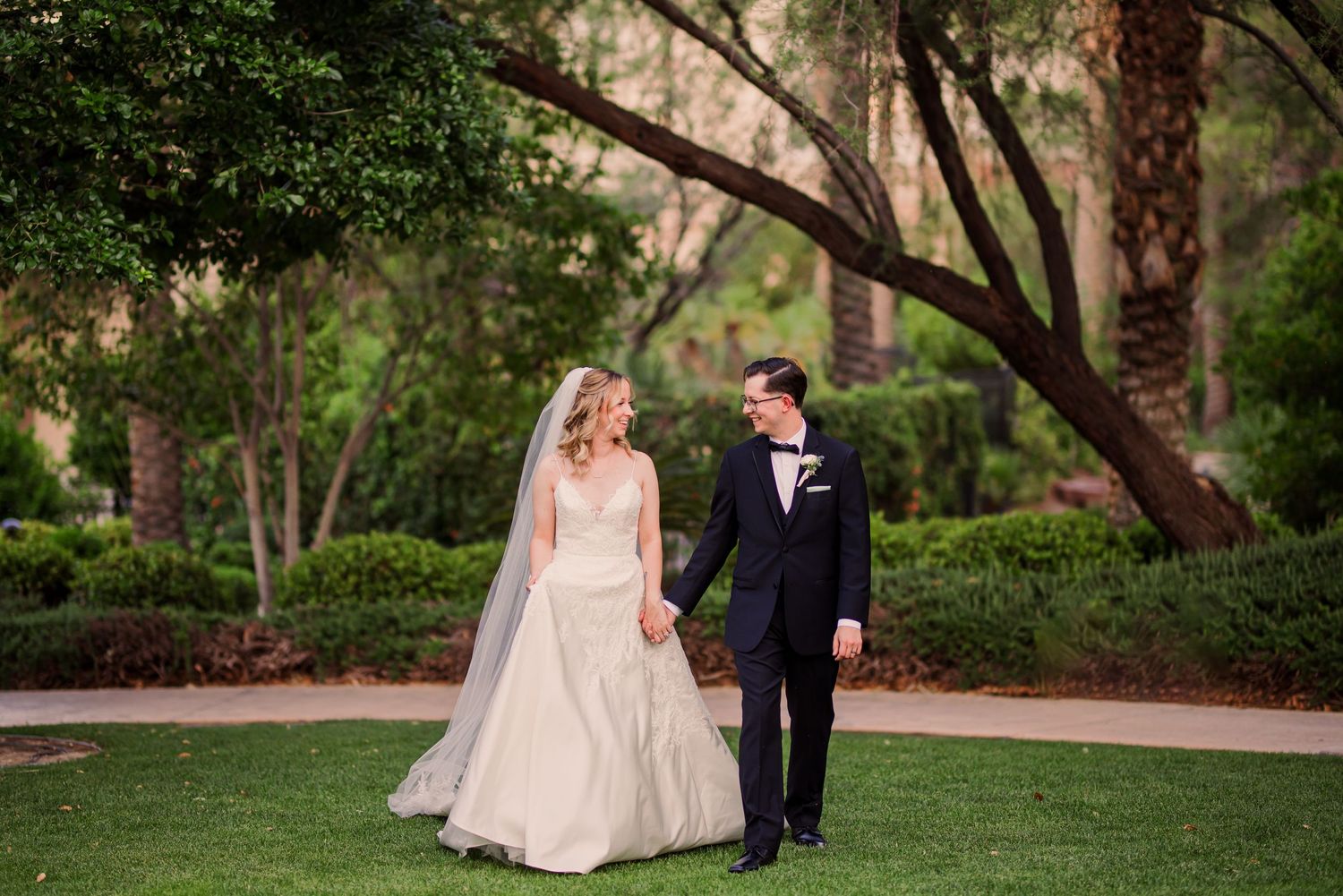 A couple in formal wedding attire walks together through a lush garden path surrounded by trees.