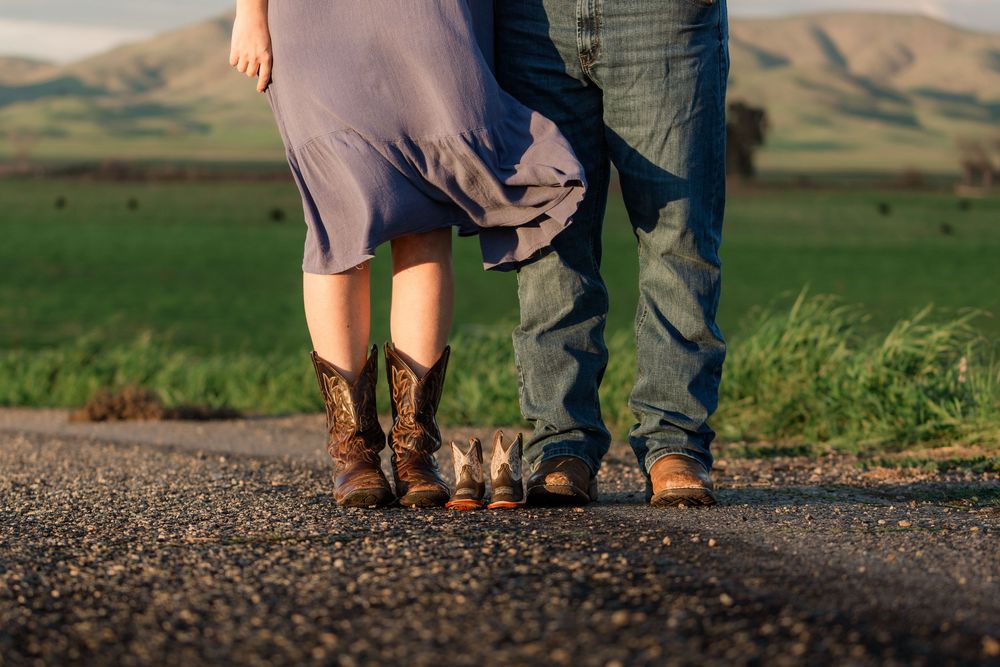Feet of a couple standing together on a dirt path with rolling hills in the background at sunset.