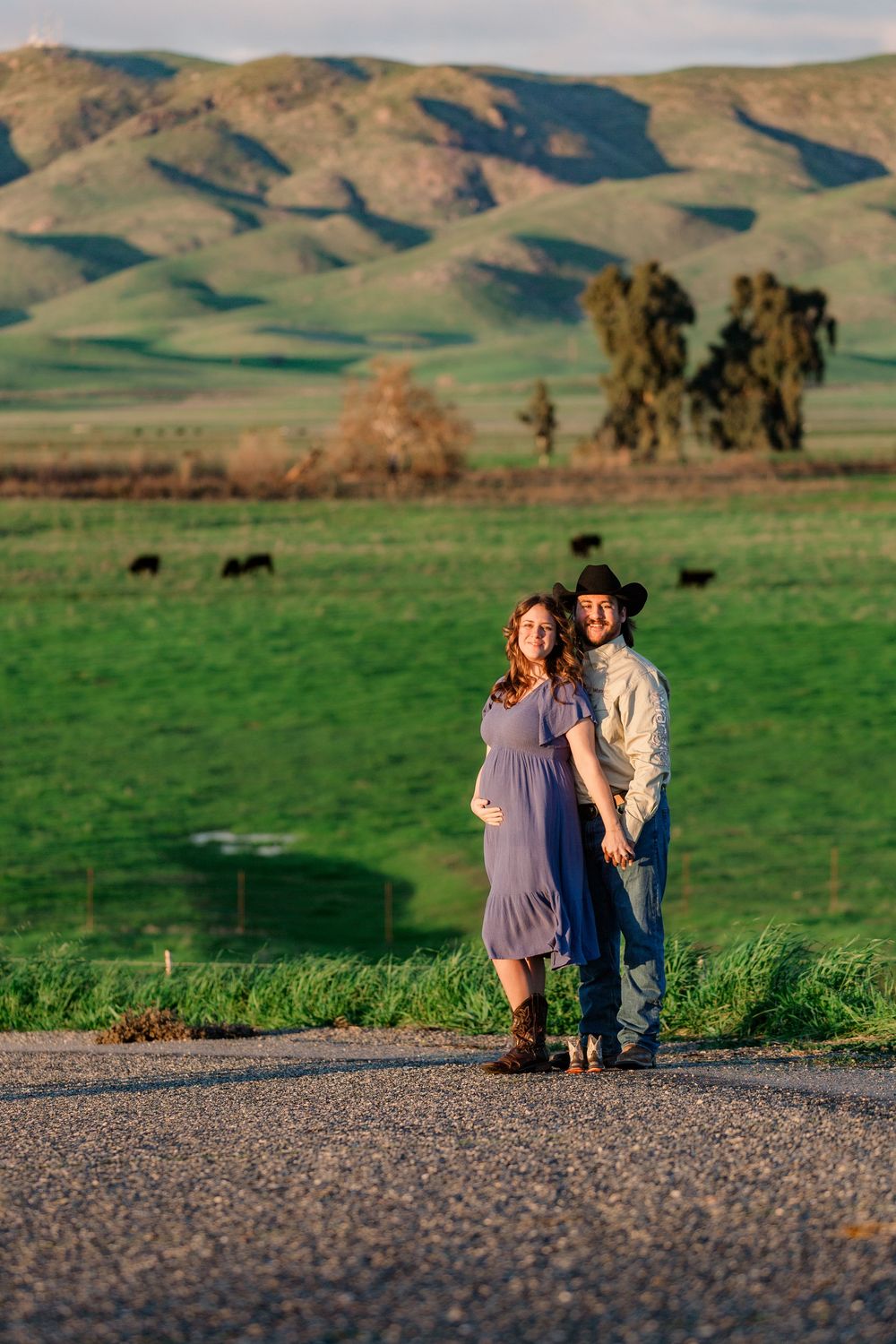 A couple sits together on a gravel road overlooking rolling green hills and mountains in a scenic rural landscape.
