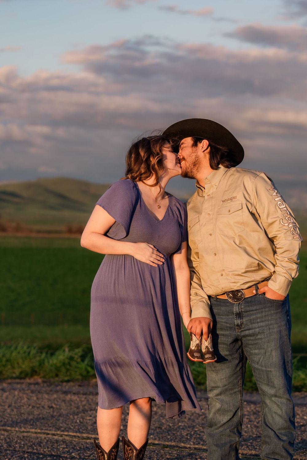A couple in a purple dress and casual attire share an intimate moment at sunset in a rural field setting.