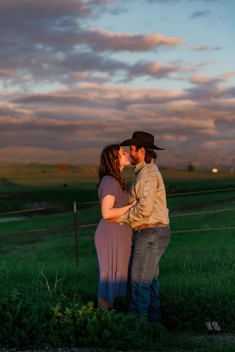 A romantic couple shares a tender moment at sunset in a rural pasture with golden clouds overhead.