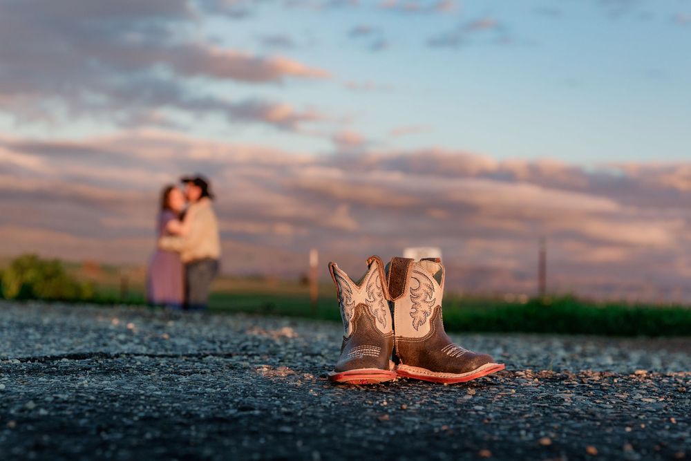 Brown cowboy boots on asphalt with a couple silhouetted against a colorful sunset sky in the background.