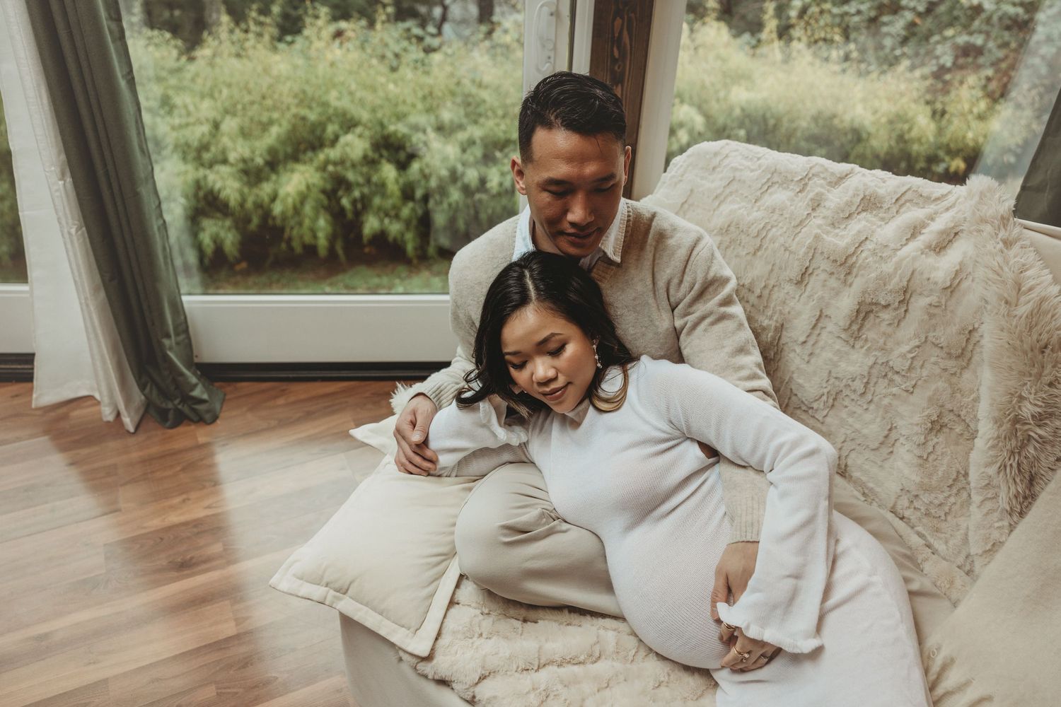 A couple relaxes together on a cozy beige couch near large windows with natural light streaming in.
