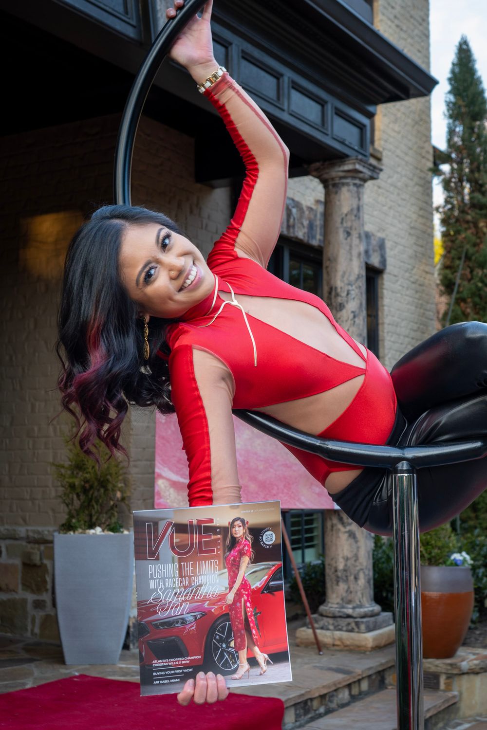 Woman in red athletic wear performs graceful pole dance moves in outdoor setting with brick building backdrop.