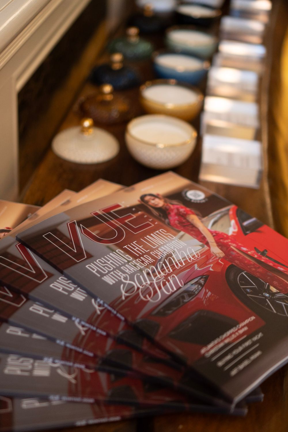 Stack of glossy magazines with red cover design displayed alongside white candles on a wooden surface.