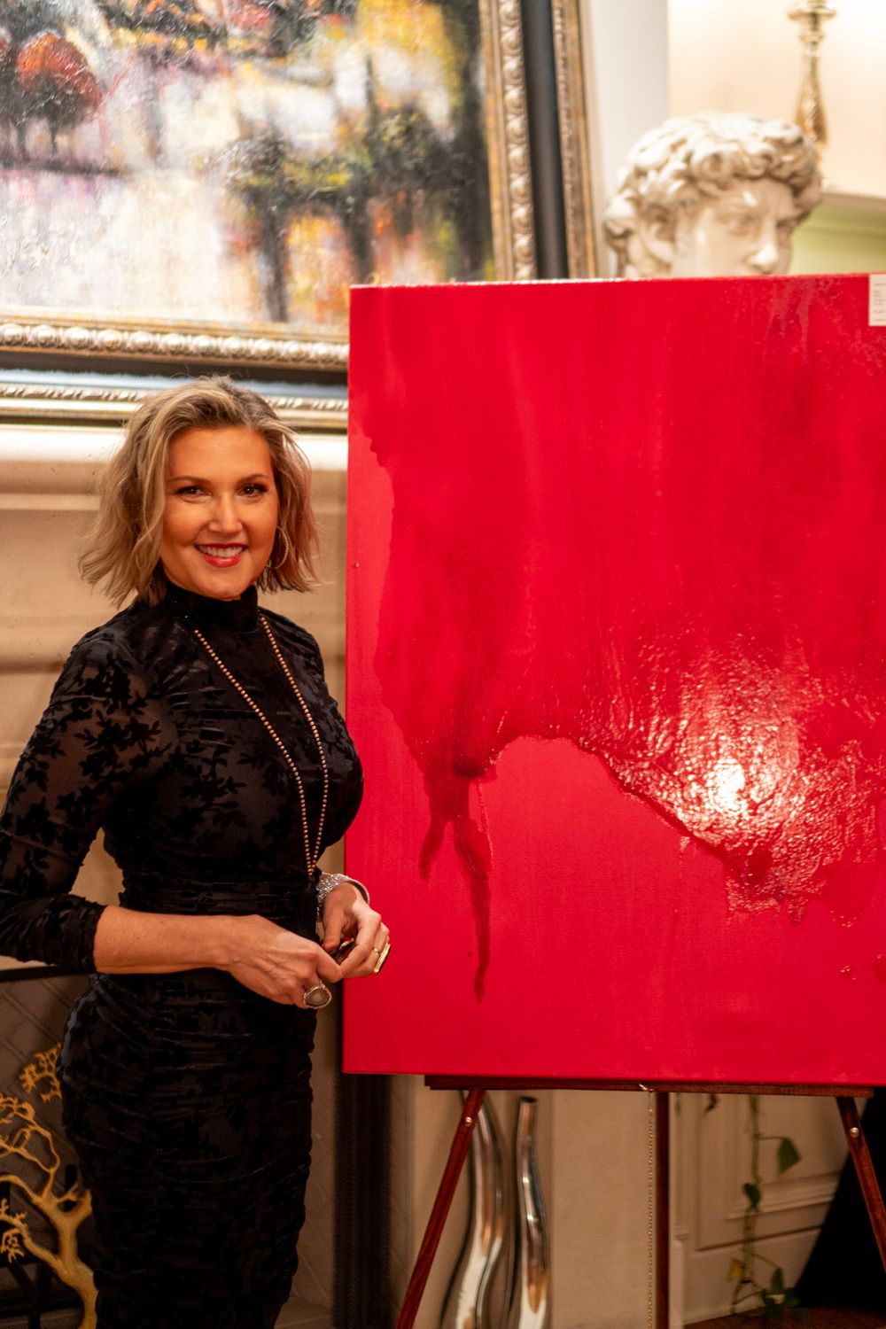Artist in black attire works on vibrant red canvas in art studio with paintings displayed in background.