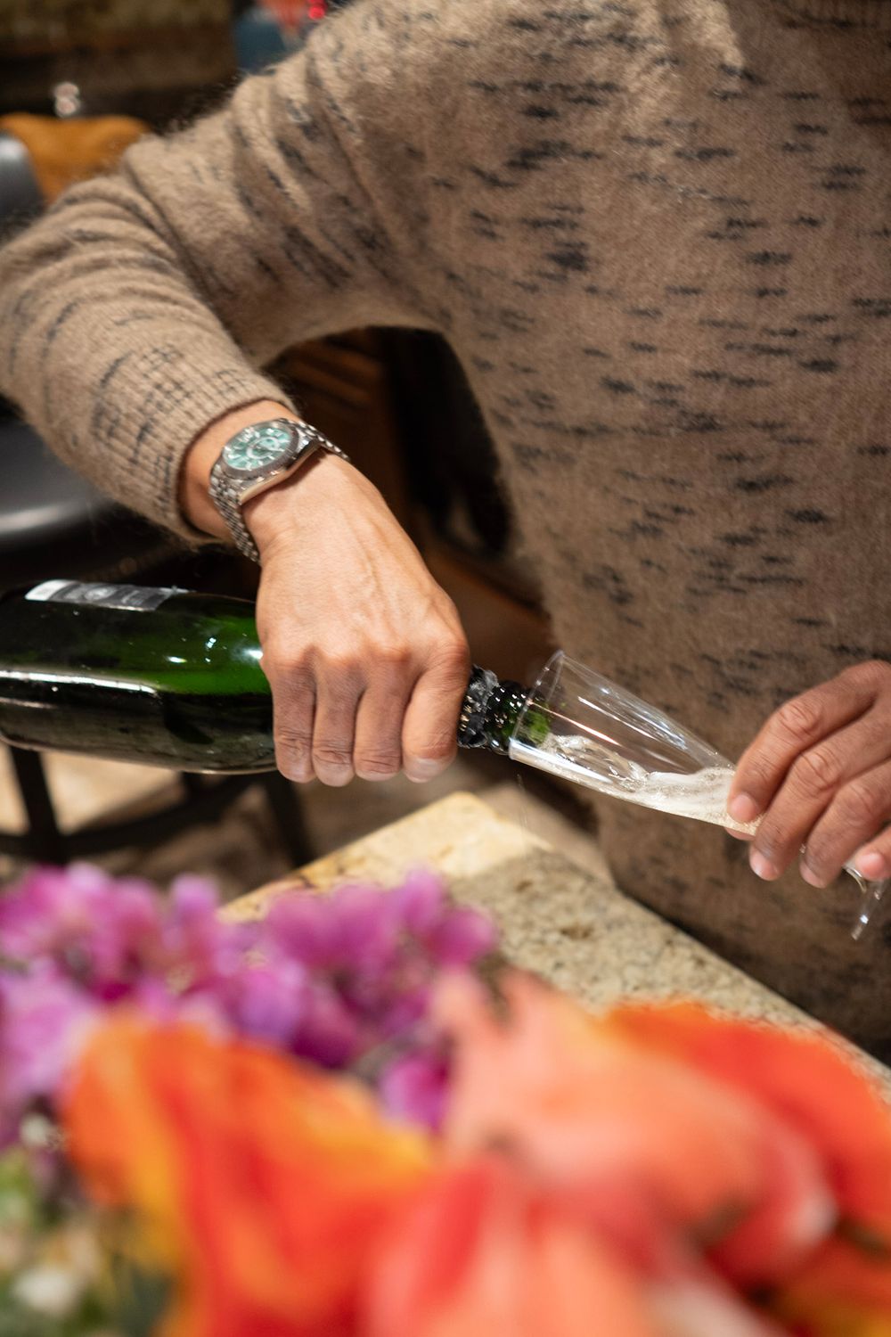 A hand wearing a decorative ring pours wine into a glass against a background of colorful flowers.