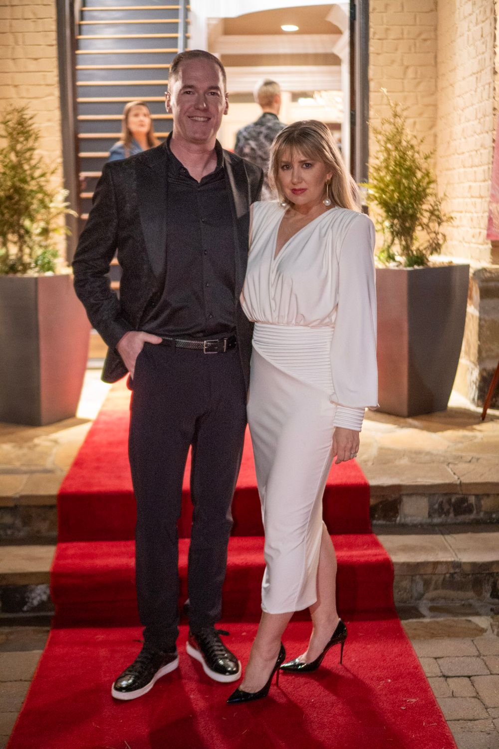 A couple poses on a red carpet outside an entrance, with the man in dark attire and woman in white formal dress.