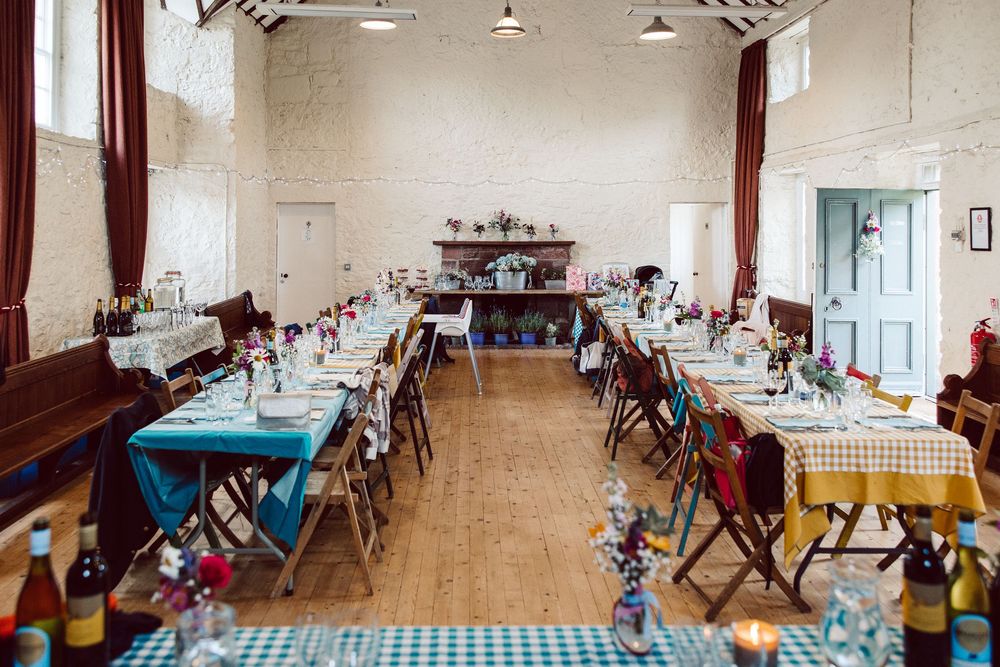 Long banquet tables with blue checkered tablecloths set up in a rustic event space with white walls and wooden floors.