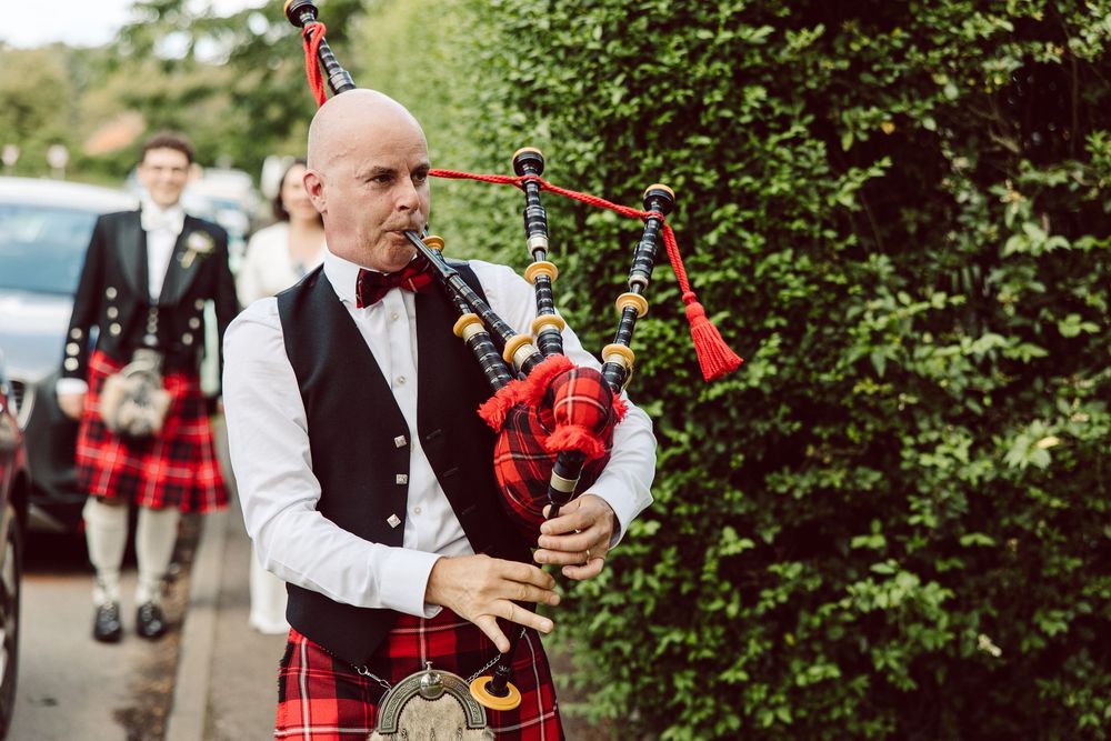 Bagpiper in traditional Scottish attire performs at outdoor event with trees in background.