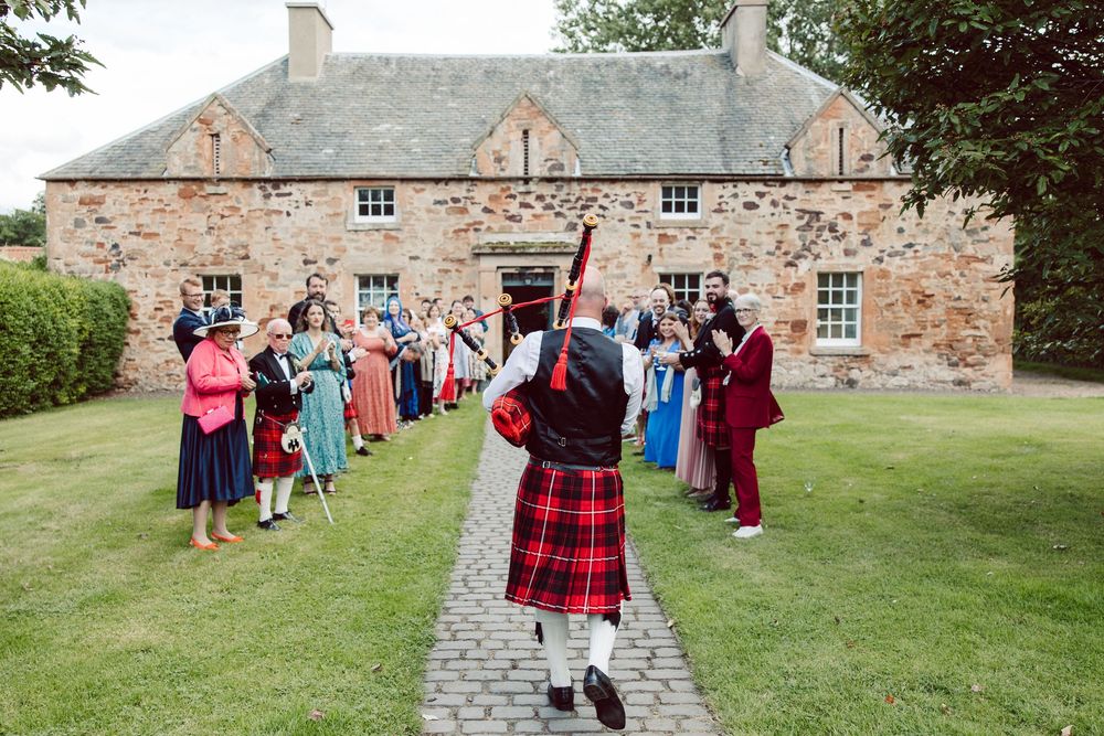 Traditional Scottish celebration with bagpiper in tartan kilt leading guests outside historic stone building.