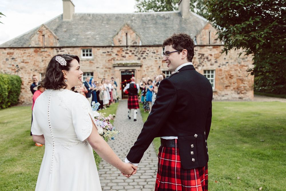 Wedding guests celebrate with champagne toasts on a sunny lawn outside a rustic brick building.