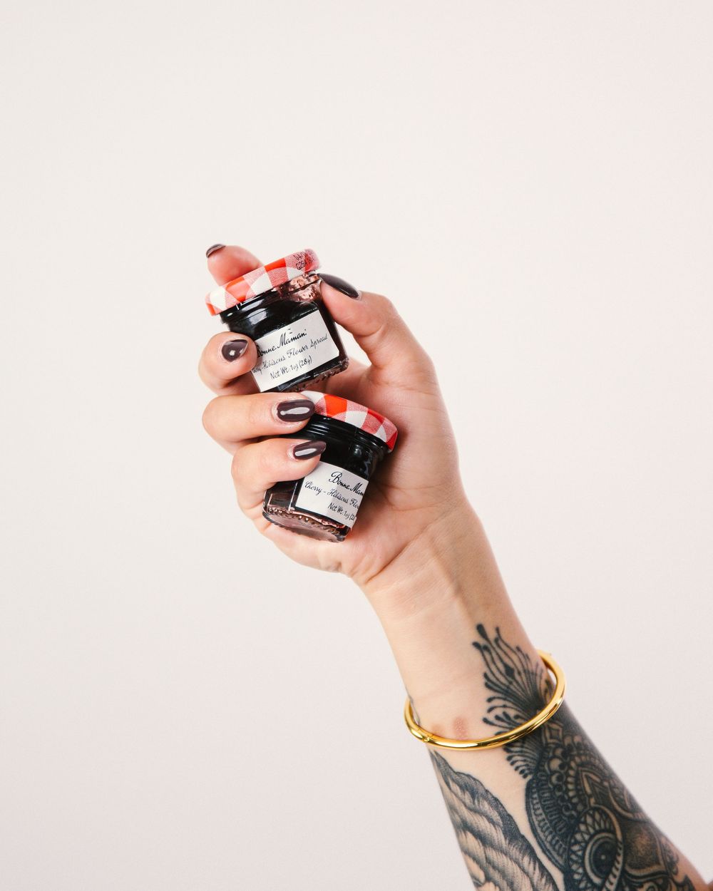 Tattooed hand holding two small glass jars with red lids against white background.