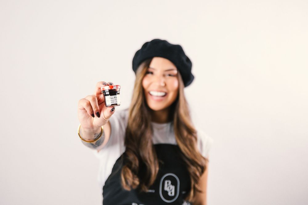 Woman in black beanie and sweater holds up small glass bottle while smiling at camera.