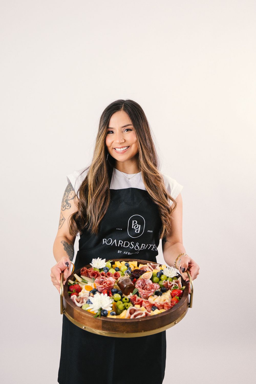 A person in a black t-shirt holding a large wooden platter filled with colorful charcuterie and fresh ingredients.